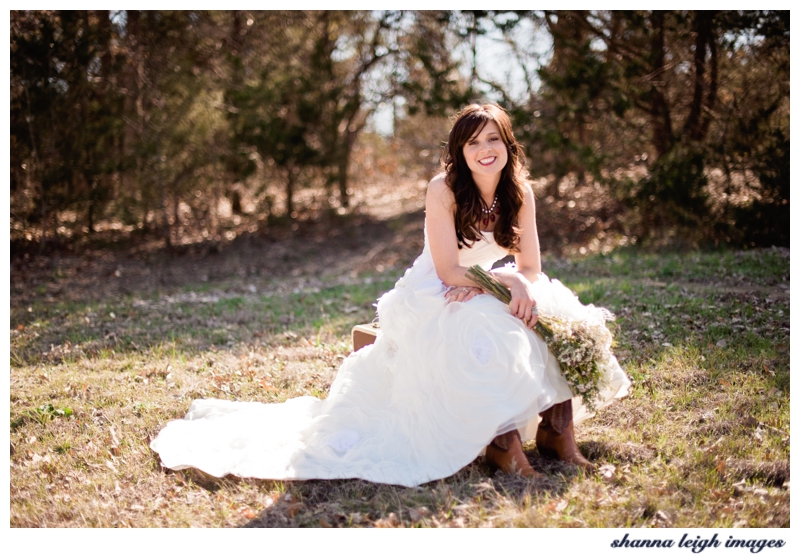 Beautiful bride Emily in her custom wedding dress by Bliss Bridal of Fort Worth, Texas in a field with her gorgeous statement necklace and cowboy boots holding a bouquet of wild flowers.